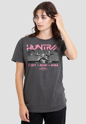 Young woman wearing dark gray T-shirt featuring three animated female figures in action poses with text "HUNTRX," "Zoey Rumi Mira," and "K-Pop Demon Hunters."