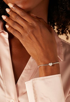 Zilveren armband met een hartbedel, met een gestructureerd oppervlak. Draagt op een hand, met een roze satijnen shirt gedeeltelijk zichtbaar.
