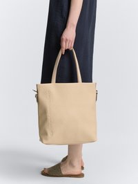 Hand holding beige tote bag with embossed logo, wearing black textured dress and woven slide sandals, standing against plain background.