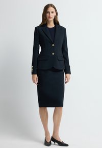 Granatowa marynarka ze złotymi guzikami, z klapami o wykroju, oraz przednimi kieszeniami, w połączeniu z ołówkową spódnicą do kolan i czarnymi szpilkami.