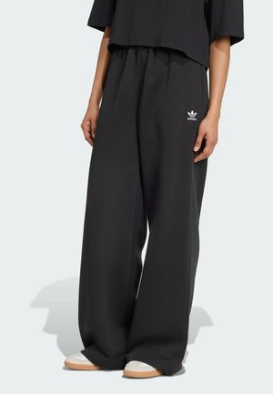 Henkilö, jolla on päällään löysät mustat Adidas-housut, joissa on joustava vyötärö ja logo vasemmassa reidessä, yhdistettynä valkoisiin lenkkitossuihin, joissa on beige pohja.