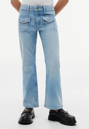 Person iført lyseblå jeans med to store knappede forlommer, kombineret med sorte loafers med guldhardware.