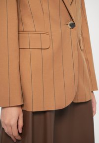 Hnedý prúžkovaný blazer s jedným gombíkom, vreckom na prsiach a ostrými klopami. Tkanina vyzerá štruktúrovaná a hladká.