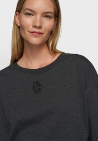 Camiseta de gran tamaño en gris carbón con una textura suave y un escote amplio. Presenta un discreto logo negro bordado en el pecho.