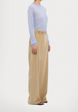 Kobieta w jasnoniebieskim swetrze z długim rękawem, wpuszczonym w beżowe, szersze spodnie z wysokim stanem wykonane z sztruksu, nosząca czarne sandały z odkrytymi palcami.