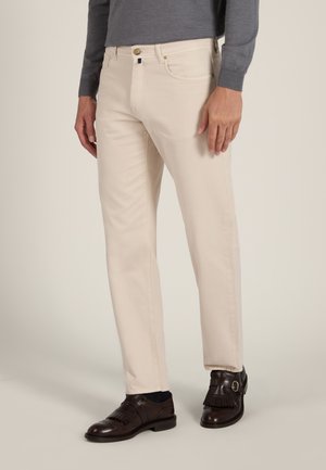 Mann trägt beige gerade geschnittene Hose, dunkelbraune Lederschuhe mit Fransen und ein graues Langarmhemd vor einfarbigem Hintergrund.