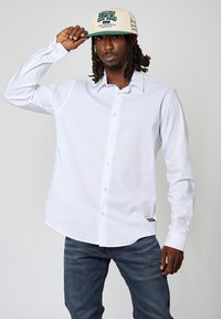 Witte langemouwen shirt met een button-down ontwerp, eenvoudige snit en subtiel logo. Draagt een beige pet met groene tekst.