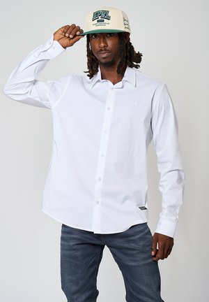 Witte langemouwen shirt met een button-down ontwerp, eenvoudige snit en subtiel logo. Draagt een beige pet met groene tekst.