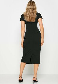 Robe midi noire ajustée avec manches courtes, fente à l'arrière, texture lisse et design moulant. Associée à des sandales à talons hauts noires.