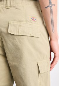 Beiga cargobyxor i bomull med fickor med lock, detaljerad sömnad och en logotagg på bakfickan. Slät, matt yta.