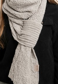 Lichtgrijze gebreide sjaal met textuur en ribbelstructuren, voorzien van een klein label en losjes over een zwarte trui gedrapeerd.