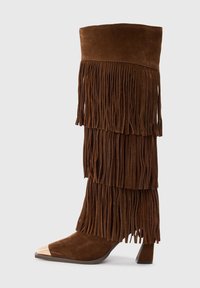 Stivale marrone in suede alto fino al ginocchio con frange stratificate e una punta metallica dorata su un tacco basso.