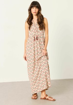 Woman with long brown hair wearing a sleeveless floral maxi dress and brown sandals standing against a plain wall.