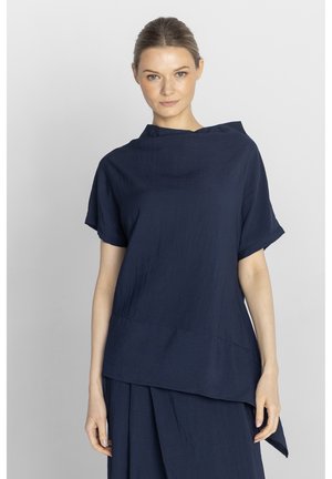 Kobieta o jasnej karnacji i związanych do tyłu włosach, ubrana w luźną, ciemnogranatową bluzkę z krótkim rękawem oraz pasującą spódnicę na jednolitym tle.