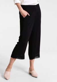 Svarta korta byxor med avslappnad passform, med sidofickor och spetsdetalj vid fållen. Materialet verkar lätt och mjukt.