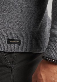 Nahaufnahme eines grauen Strickpullovers mit einem Superdry-Label, kombiniert mit dunklen Hosen und einem Perlenarmband am Handgelenk.