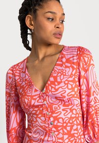 Vestido de manga longa com um padrão vibrante em rosa e vermelho, apresentando um decote profundo, frente franzida e detalhes em botão. Textura de tecido suave.