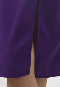 Fustă violet cu o textură fină, cu o fanta laterală care dezvăluie pielea. Linia de jos este dreaptă, cu detalii de cusătură îngrijite.