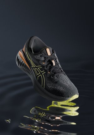 Chaussure de running ASICS noire et jaune néon avec lacets, flottant au-dessus d'une surface noire réfléchissante ondulée.