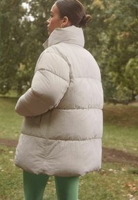 Lys grå pufferjakke med høj krave, der har et quiltet design og en let blank tekstur, sammen med grønne leggings.