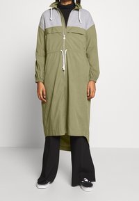 Chaqueta larga verde oliva con detalles grises, cintura con cordón, cremallera frontal y puños elásticos. Combinada con pantalones y zapatos negros.