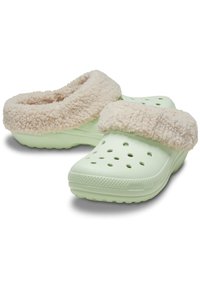 Zuecos de color verde claro con una suela texturizada, forma redondeada y un suave forro de piel sintética beige alrededor del tobillo. Orificios de ventilación a los lados.