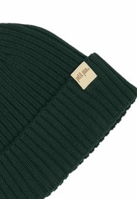 Bonnet tricoté vert foncé avec texture côtelée, arborant une étiquette rectangulaire beige avec le nom de la marque brodé sur le revers.