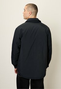 Black lightweight jacket with a relaxed fit, long sleeves, and a high collar. Smooth texture with a slightly glossy finish, tailored back hem.