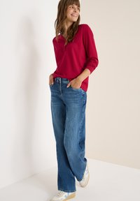 Rød langærmet top med knapdetalje, parret med brede blå jeans og hvide sneakers med lyseblå accenter på en neutral baggrund.