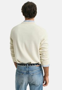 Crème-kleurige trui met lange mouwen, ronde hals en gebreide zoom, gecombineerd met blauwe denim jeans en een patroonstrik.