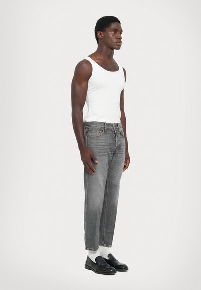 Young man wearing white tank top, gray jeans, white socks, and black loafers standing against a plain light background.