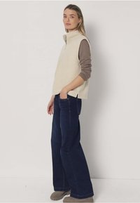 Beige Fleeceweste mit Reißverschluss, kombiniert mit einem braunen Langarmshirt, dunkelblauen Jeans mit weitem Bein und grauen Stiefeln.