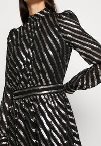 Robe noire avec des rayures verticales argentées, des manches longues bouffantes, un col haut et une taille ajustée. Tissu texturé avec une finition scintillante.