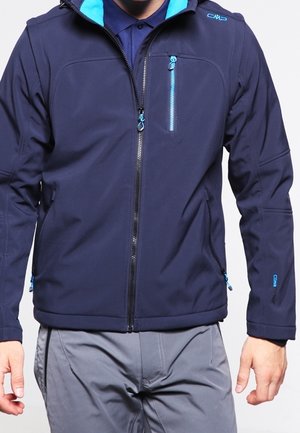 Homme portant une veste zippée bleu marine avec des accents de fermeture éclair bleu clair et un pantalon gris, debout devant un fond uni.