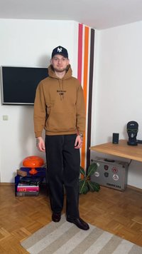 Καφέ φούτερ με κουκούλα, μπροστινή τσέπη και λογότυπο, μαύρο παντελόνι με φαρδιά μπατζάκια και καφέ παπούτσια. Το φόντο περιλαμβάνει έναν τοίχο με πολύχρωμες ρίγες.