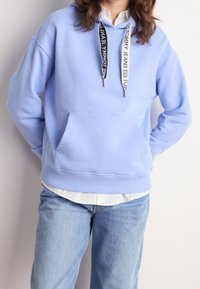 Person som bär en ljusblå hoodie med svarta och vita dragkedjor från Tommy Jeans, tillsammans med ljusblå jeans och en vit skjorta under.