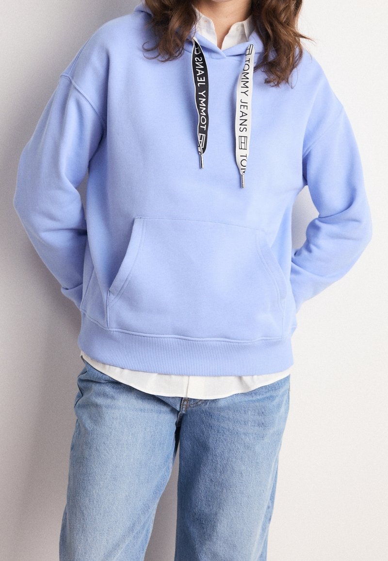 Person som bär en ljusblå hoodie med svarta och vita dragkedjor från Tommy Jeans, tillsammans med ljusblå jeans och en vit skjorta under.