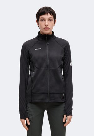 Ung person med kort mørkt hår iført en sort Mammut lynlåstrøje og mørke bukser, stående mod en ensfarvet lys baggrund.
