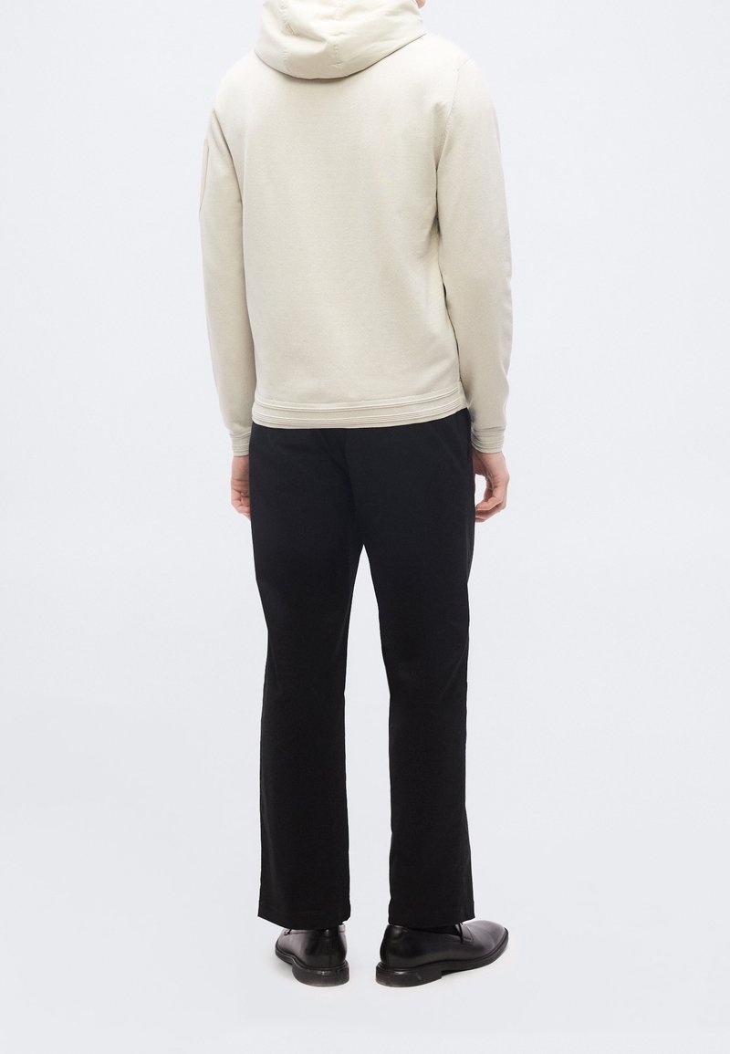 Personne portant un sweat à capuche beige clair, un pantalon noir et des chaussures noires, debout de dos sur un fond blanc uni.
