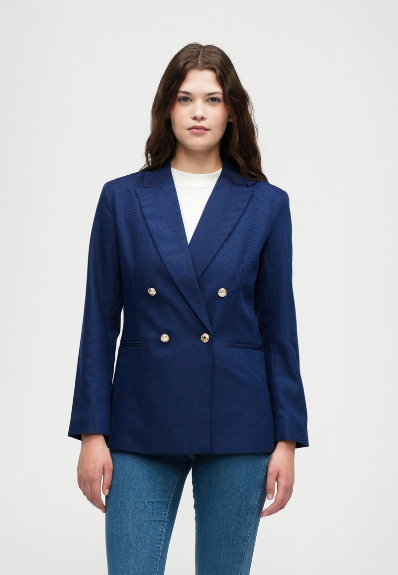 Marineblauwe dubbelgeknoopte blazer van een gestructureerde stof, met gouden knopen, geknipte revers en twee verborgen zakken. Gecombineerd met blauwe jeans.