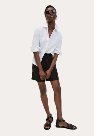 Hvid skjorte med opsmøgte ærmer, sorte shorts og sorte sandaler. Model iført solbriller står foran en neutral baggrund.