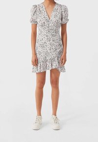 Kvinna står klädd i en kort vit blommig klänning med puffärmar och vita sneakers på en enfärgad bakgrund.