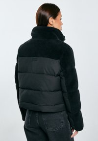 Černá péřová bunda s vysokým límcem, vyrobená z měkkého materiálu v horní části a hladké tkaniny dole, navržená v boxovém siluetě.