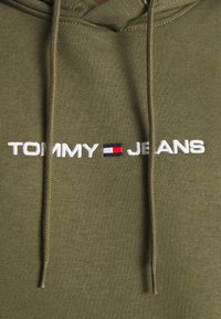 Sudadera color verde oliva hecha de una mezcla de algodón, con un logo frontal que presenta el bordado "TOMMY JEANS" y un emblema rectangular en rojo, blanco y azul.