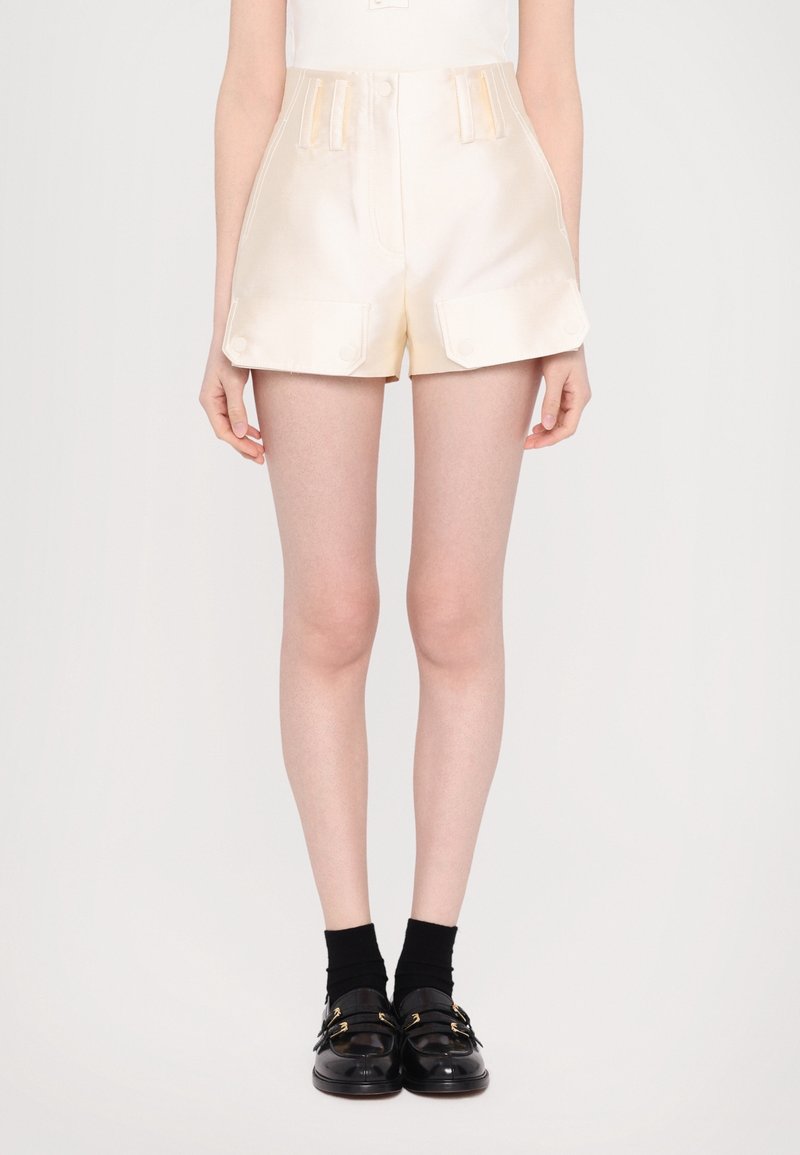 Femme portant un short crème taille haute avec poches avant à rabat, chaussettes noires et mocassins en cuir noir, debout sur fond blanc.