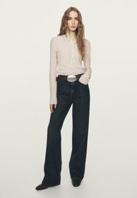 Długorękawowy, jasnoróżowy prążkowany top z trzema guzikami, zestawiony z ciemnoniebieskimi, szerokimi spodniami jeansowymi i brązowym paskiem z dużą srebrną sprzączką.