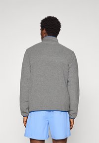 Grijze fleece jas met een hoge kraag, geribde manchetten en een blauwe voering, gedragen met lichtblauwe shorts. Zachte textuur, eenvoudig ontwerp.