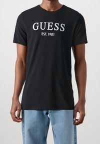 Sort bomulds T-shirt med rund hals, korte ærmer og et sølvfarvet "GUESS EST. 1981" grafik på fronten, sammen med blå denim jeans.