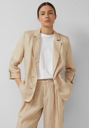 Femme portant un costume en lin beige avec manches retroussées et un t-shirt blanc, debout, les mains dans les poches devant un fond gris uni.