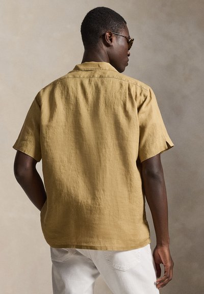 Hombre con camisa de manga corta color mostaza y pantalones blancos, de espaldas, con una mano en el bolsillo frente a un fondo neutro.